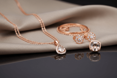 Rose gold jewelry set with necklace, ring, and earrings on a reflective surface.
